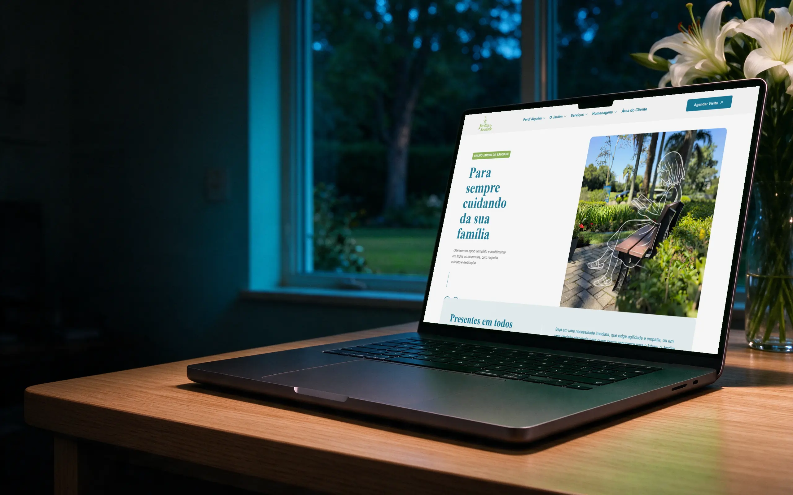 Notebook sobre a mesa de madeira clara de um escritório em casa à noite, com lírios brancos e janela para o jardim, exibindo o site do Jardim da Saudade